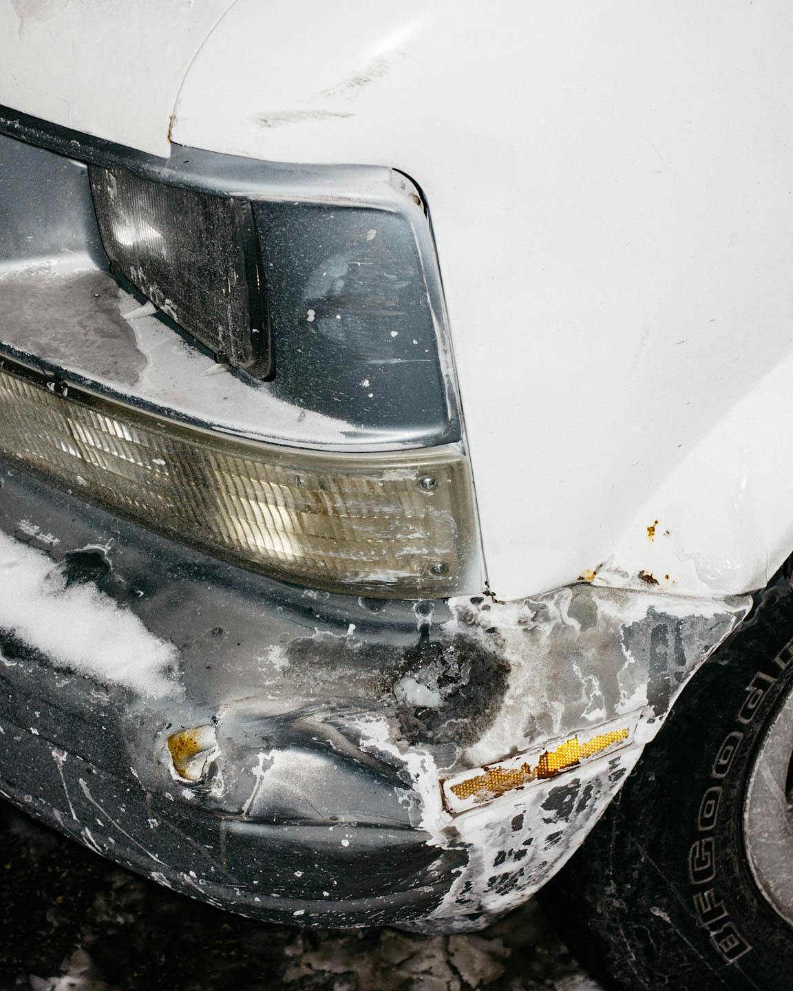 A salt-stained and snow-covered bumper of a white car.